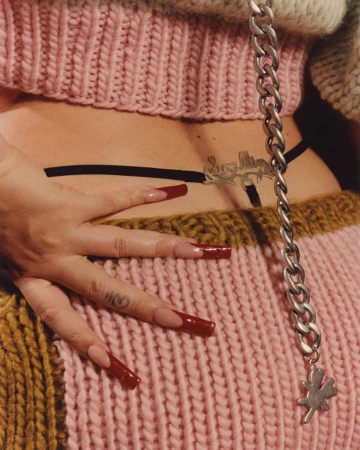 Close-up shot of a woman's lower back, hand, and long red nails, accessorized with a silver chain and a knit pink and tan sweater.