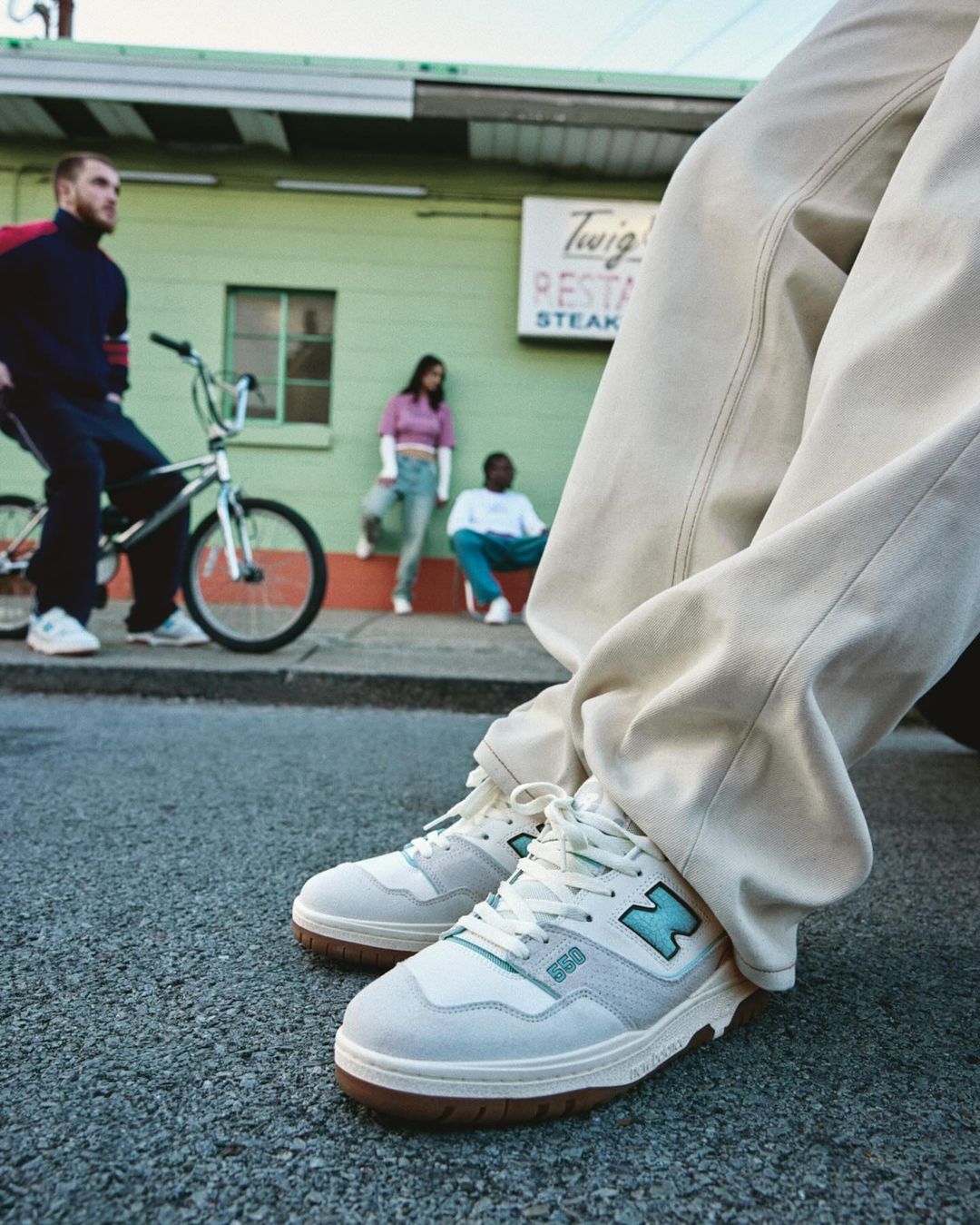 Low-angle shot of New Balance 550 sneakers on asphalt, featuring a group of young adults in the background near a green building.