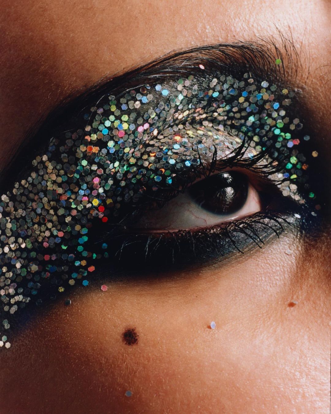 Macro close-up of a human eye with dark eyeshadow and multicolored glitter makeup on the eyelid and surrounding skin.