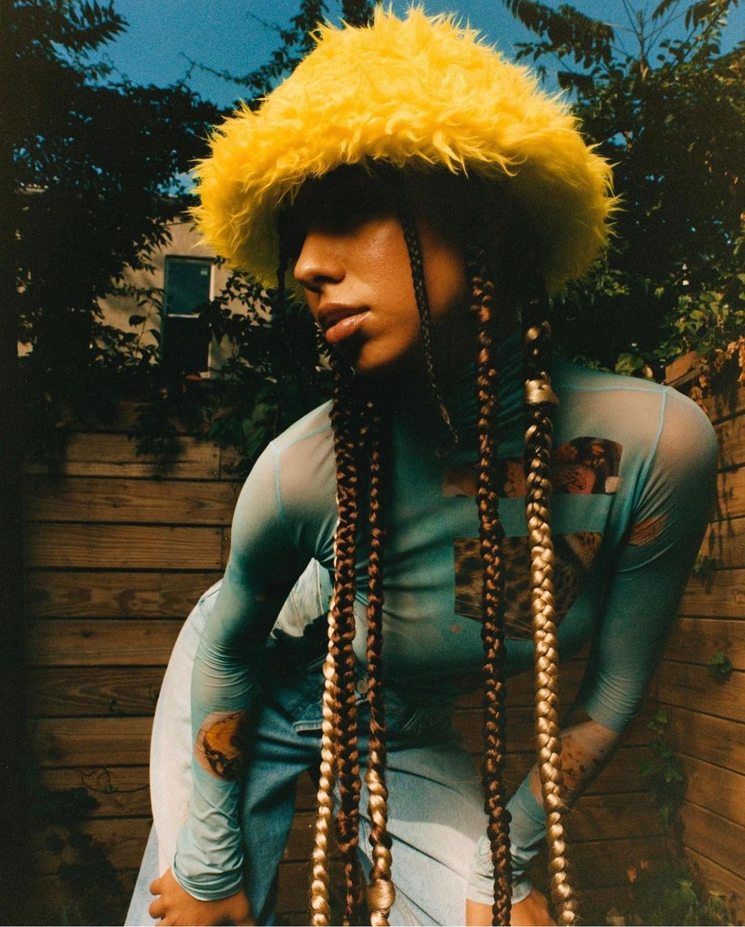 Woman in blue top and jeans poses in a yard with a wooden fence, wearing a yellow hat and long braids with decorative beads.
