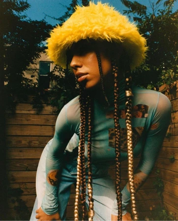 Woman in blue top and jeans poses in a yard with a wooden fence, wearing a yellow hat and long braids with decorative beads.