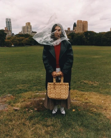 A person stands on a grassy field wearing a dark coat and skirt, with a plastic bag over their head and a patterned bag in their hands.