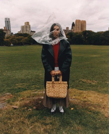 Young woman in black coat stands in a park in front of city buildings, wearing a clear plastic bag on her head and holding purse.
