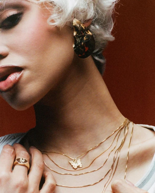 Close-up of a woman with platinum blonde hair wearing layered gold necklaces with a butterfly pendant and gold earrings.