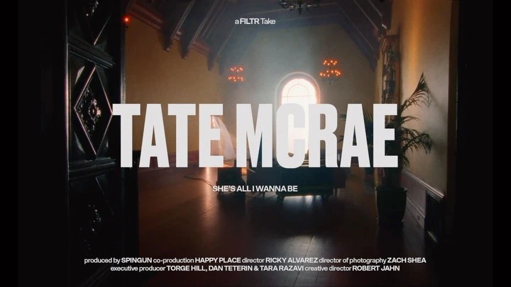Music video title card for Tate McRae's 'She's All I Wanna Be,' featuring elegant dark interior, window, potted plant.