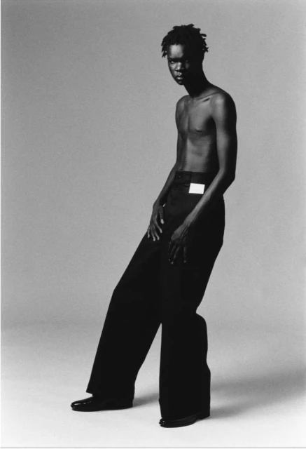 Monochrome studio portrait of an adult, athletic male model in wide-leg trousers, posing with hands, against a seamless backdrop.