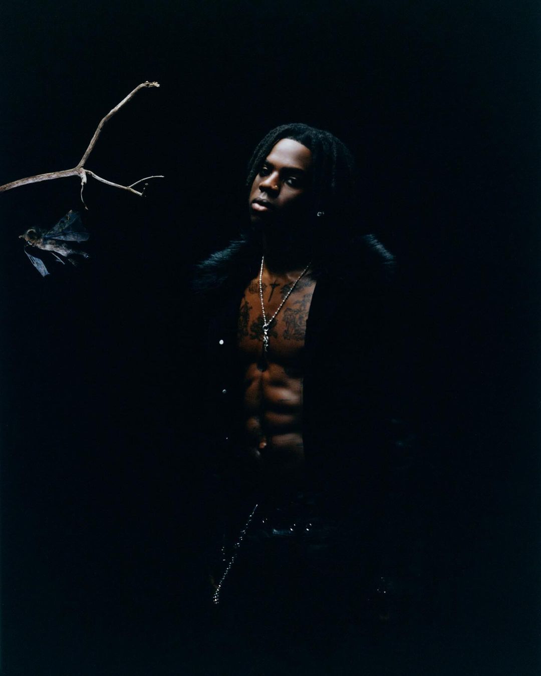 Portrait of an adult male with dreadlocks and an open black jacket posing in a dark, moody setting with a branch in the background.