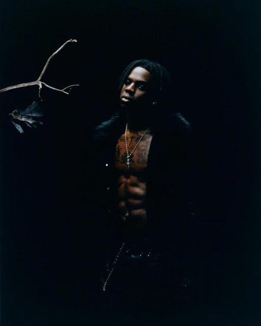 Portrait of an adult male with dreadlocks and an open black jacket posing in a dark, moody setting with a branch in the background.