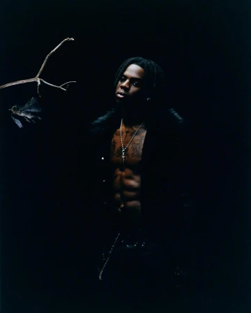 Portrait of an adult male with dreadlocks and an open black jacket posing in a dark, moody setting with a branch in the background.