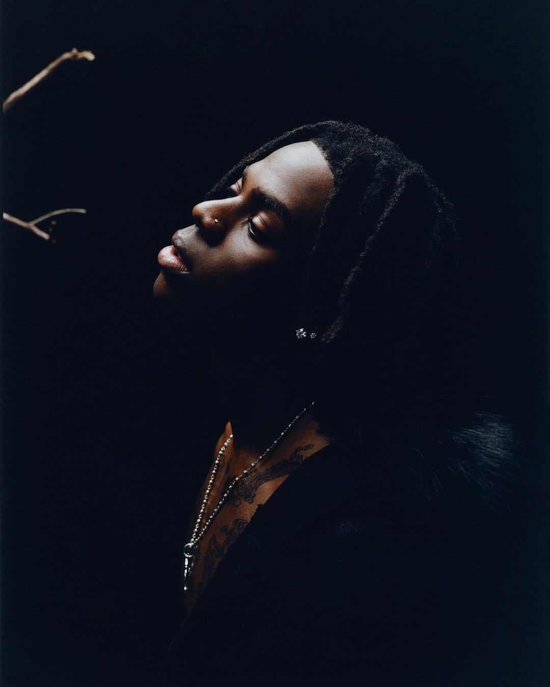 Portrait of a young Black male in a dark setting, looking up with a nose piercing and beaded necklace, against a dark backdrop.