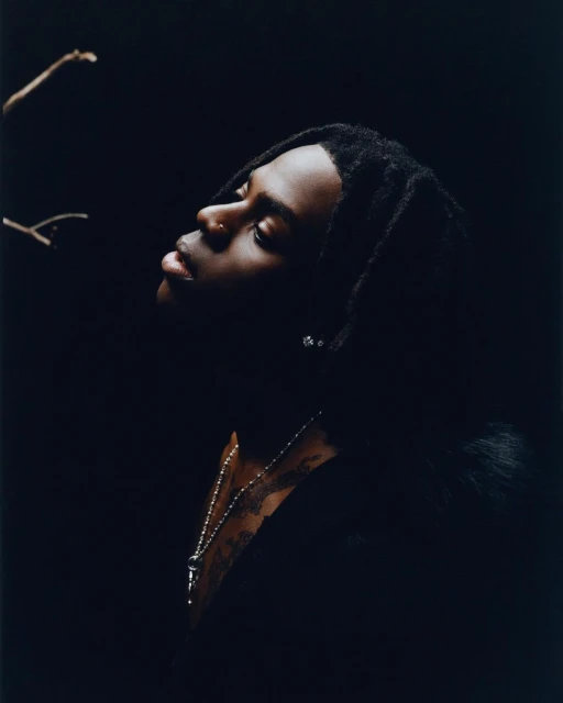 Portrait of a young Black male in a dark setting, looking up with a nose piercing and beaded necklace, against a dark backdrop.