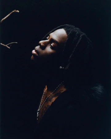 Portrait of a young Black male in a dark setting, looking up with a nose piercing and beaded necklace, against a dark backdrop.
