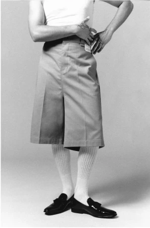 Man wearing a white tank top, pleated Bermuda shorts, ribbed socks, and loafers against a plain background