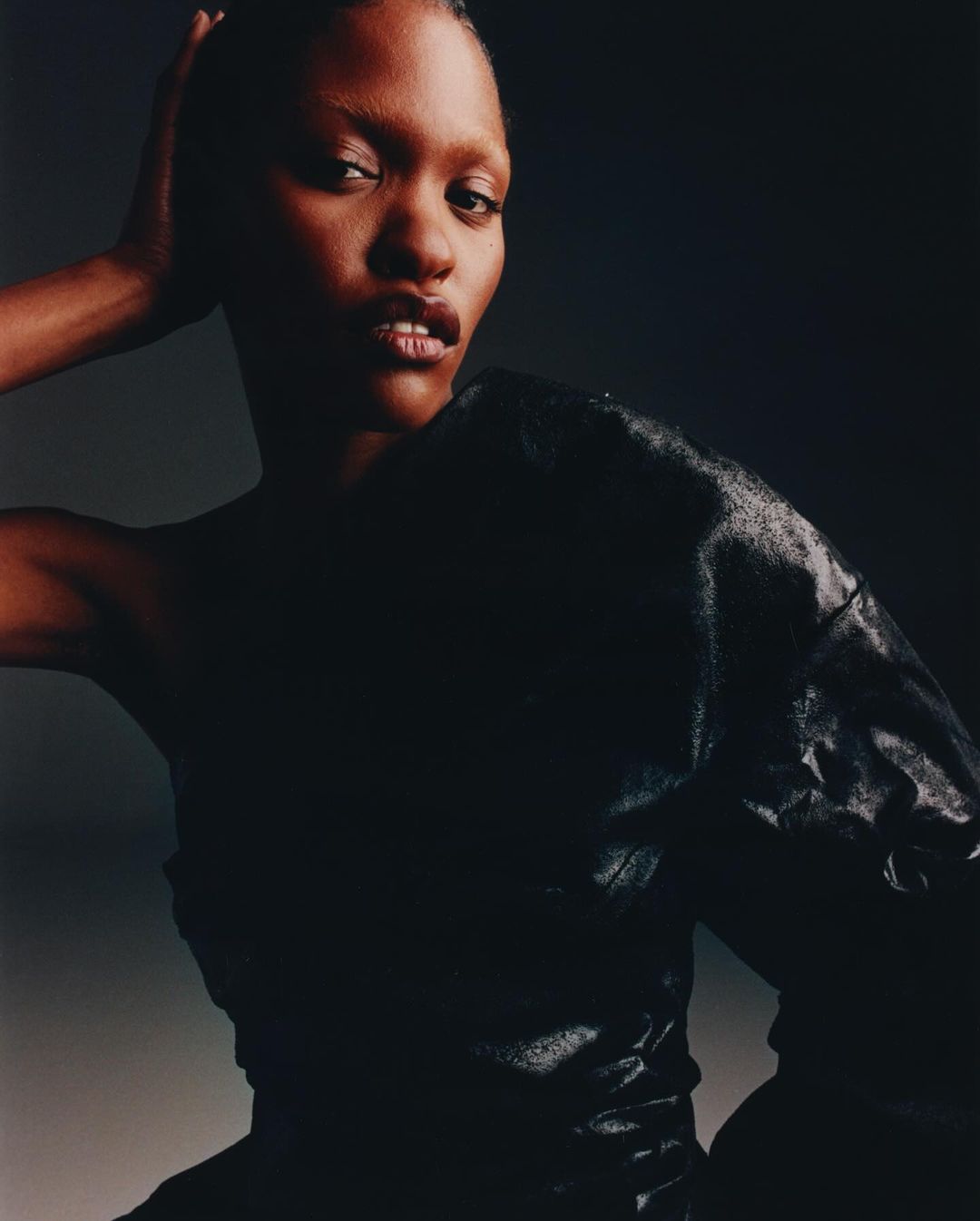 Close-up studio portrait of a black female with dark hair, wearing a black top with texture. Dark, neutral background.