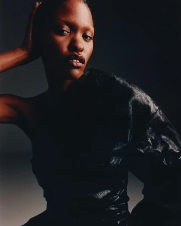 Close-up studio portrait of a black female with dark hair, wearing a black top with texture. Dark, neutral background.