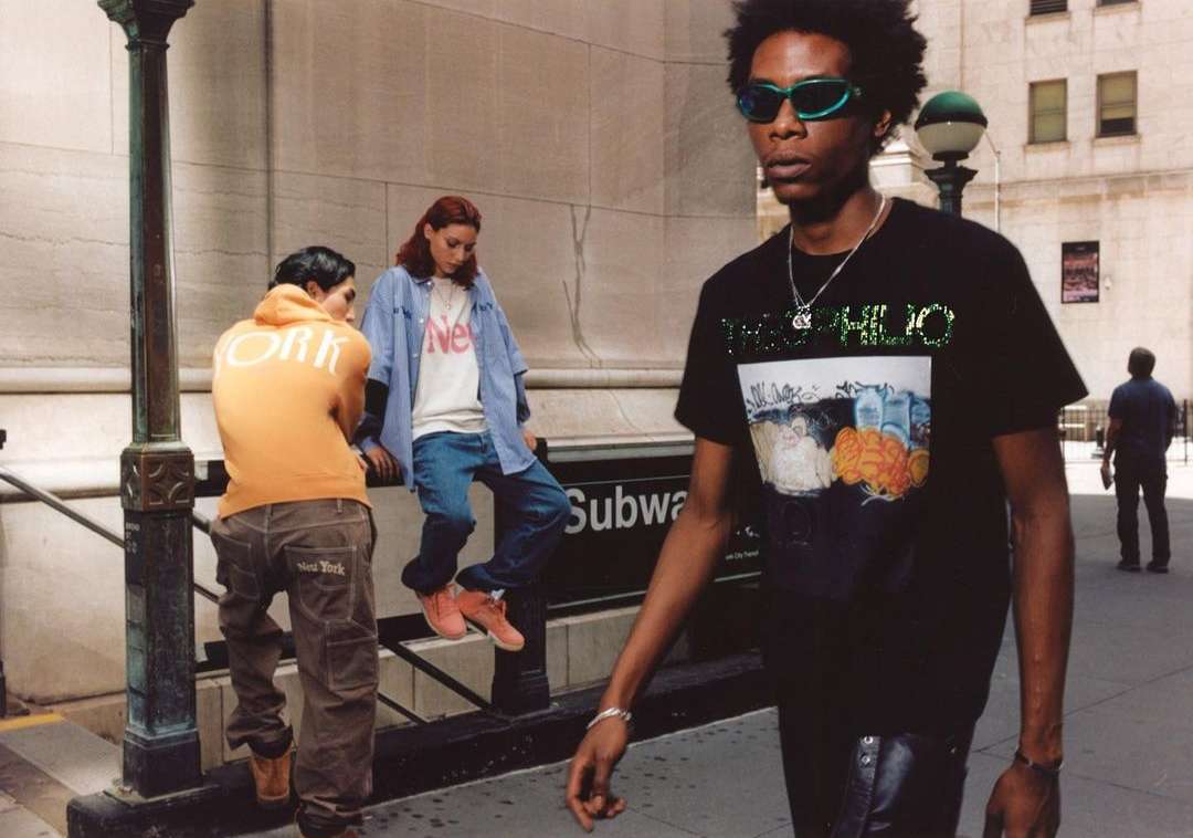 Three people in urban fashion near a subway entrance; one man in sunglasses and a graphic tee walks, while two others sit nearby.