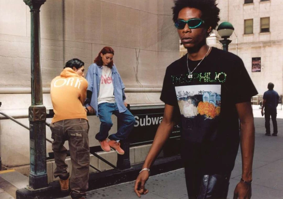 Three people in urban fashion near a subway entrance; one man in sunglasses and a graphic tee walks, while two others sit nearby.