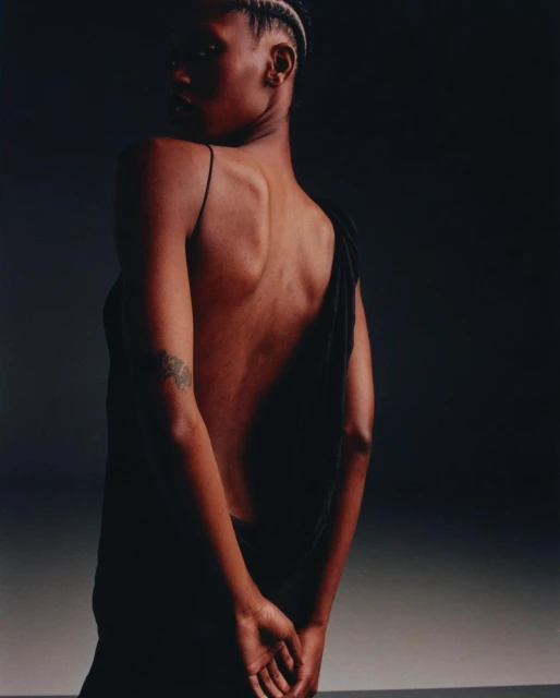 Back view of a woman with dark skin and braided hair wearing a draped black dress against a shadowy gradient backdrop, fashion shot.