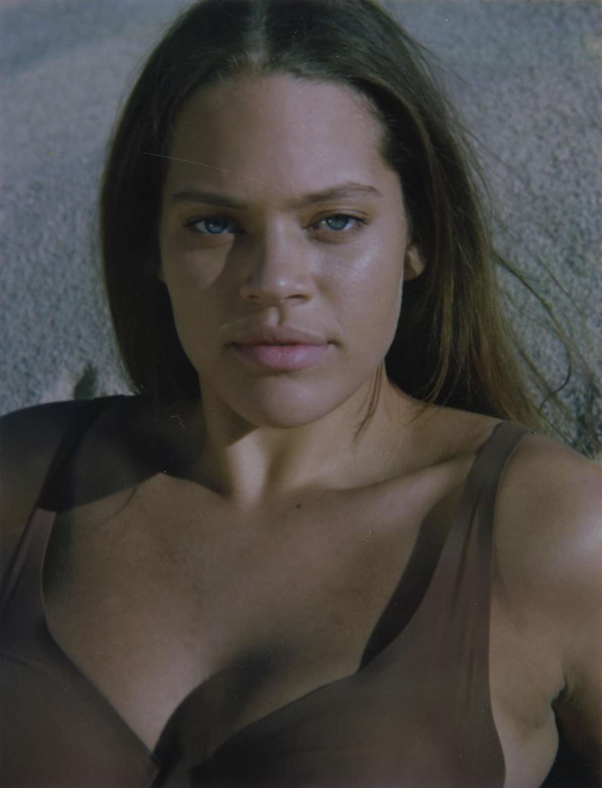 Close-up portrait of an adult female with long brown hair and blue eyes, wearing a brown top against a light gray background.