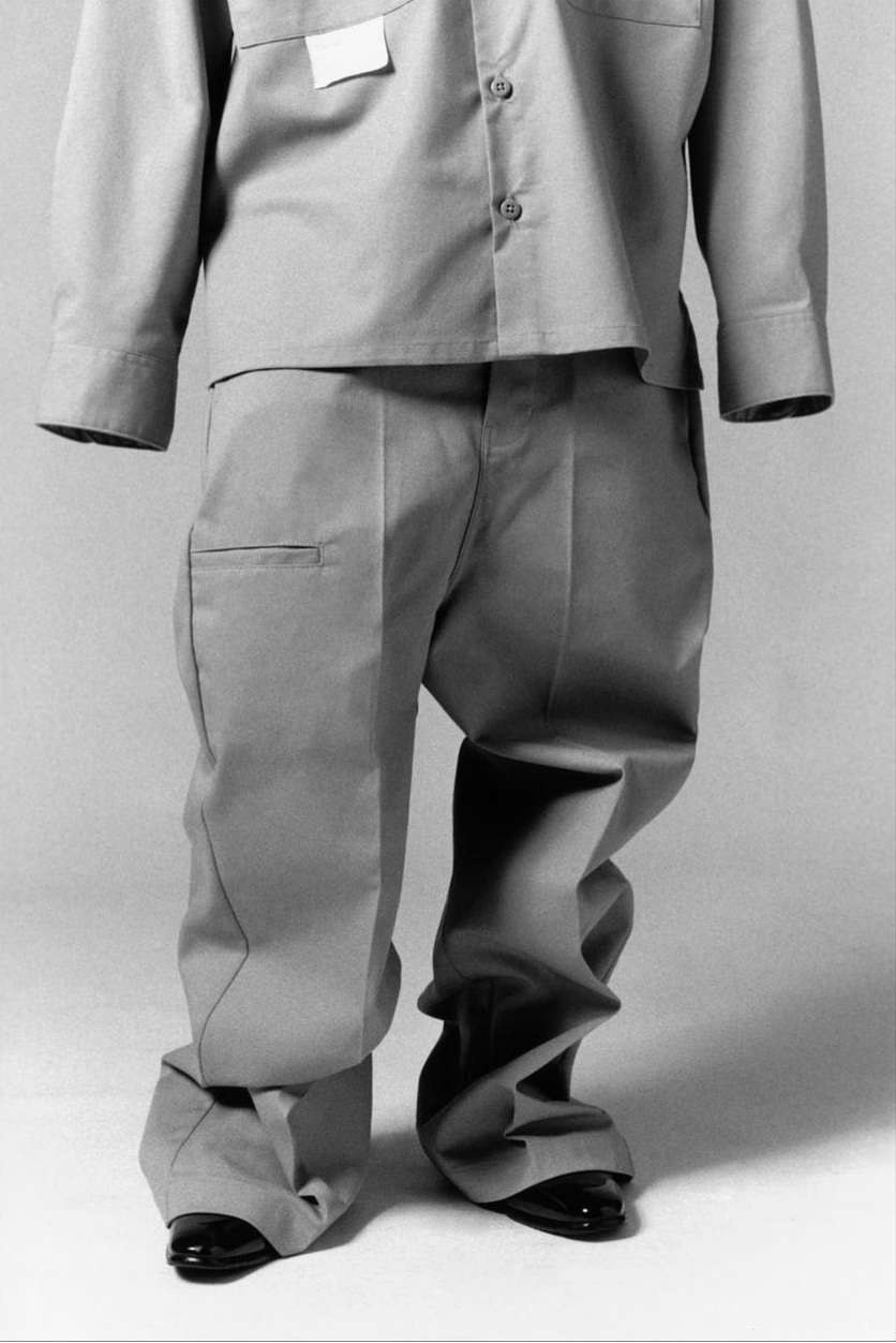 A full outfit on display features a tan, long-sleeved collared shirt, matching trousers with distorted legs, and shiny black shoes.