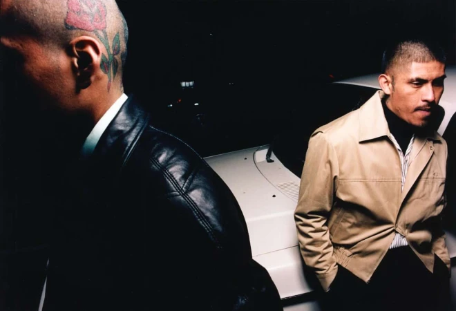 Two men near a white car at night, one wearing a black leather jacket with a rose tattoo on his head and another in a tan jacket.