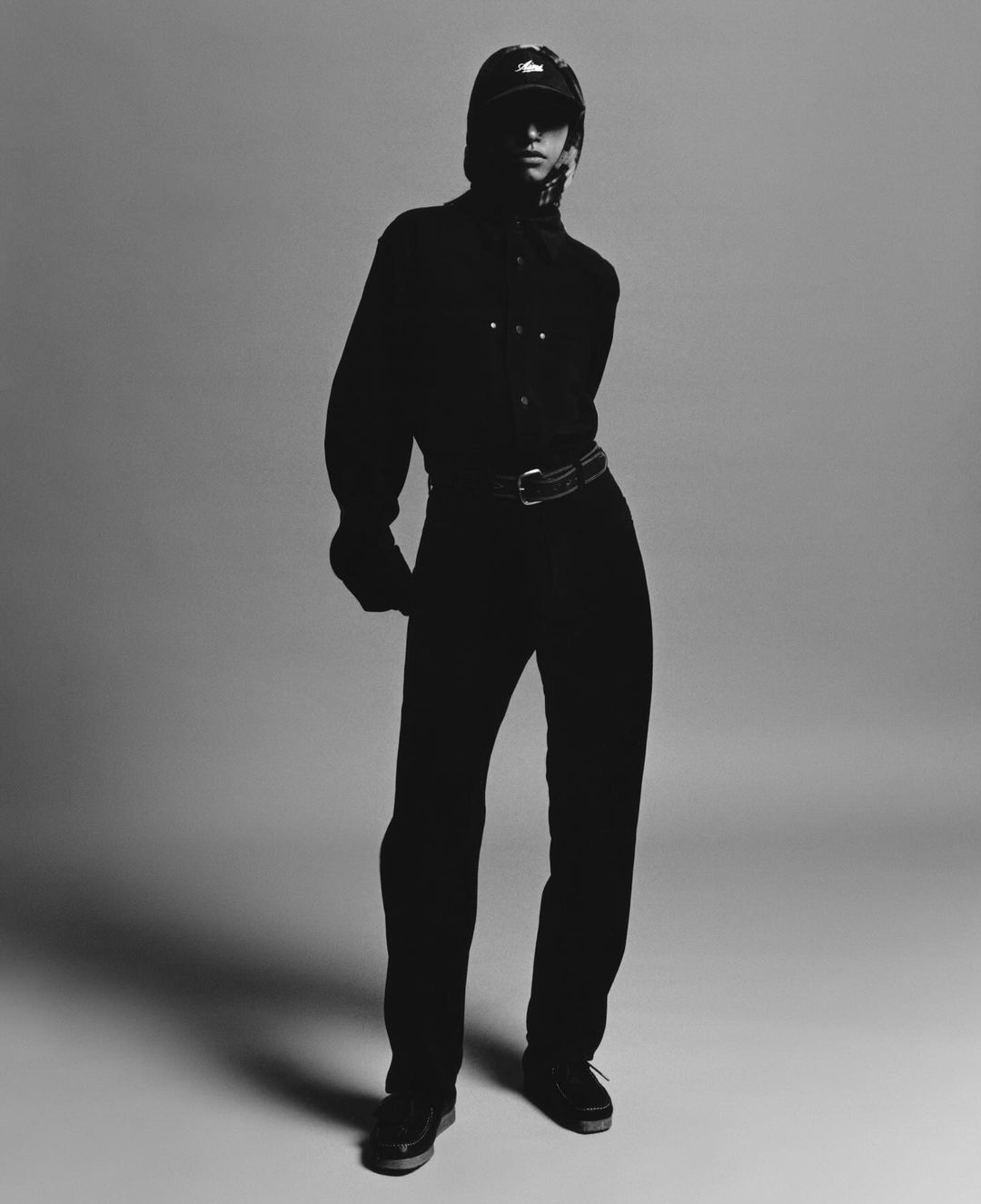Monochrome fashion portrait of an adult wearing a cap, button-down shirt, and trousers, posed in a photo studio with soft lighting.