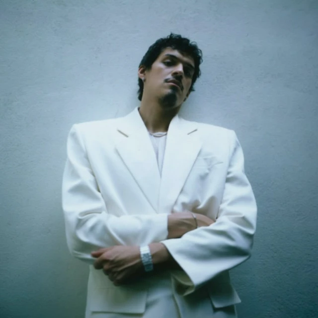 Portrait of a man in a white suit posing against a white wall, looking at the camera with arms crossed and a neutral expression.