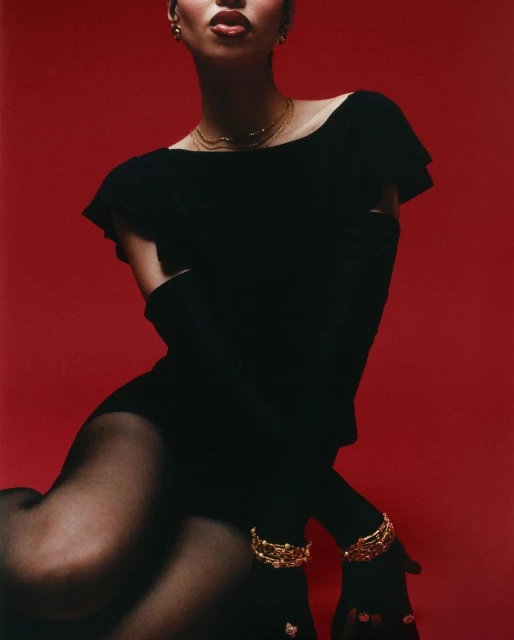 A woman in a black dress with gold bracelets poses against a red background in a fashion studio photograph.