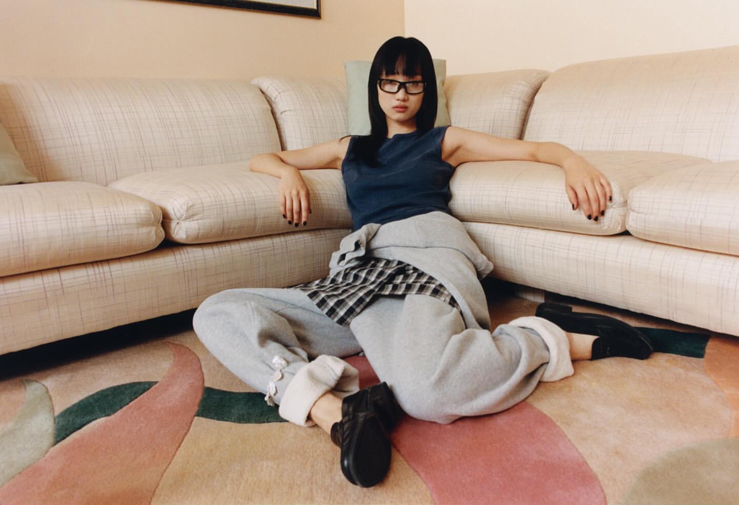 Woman in a blue tank top and gray sweatpants sits cross-legged on an area rug, posing in front of a beige couch in a bright indoor space.