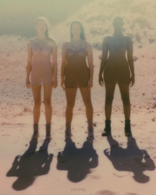 Three diverse fashion models wearing neutral-toned dresses and boots, standing in a desert landscape, with long shadows stretching forward.