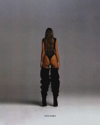 A woman facing away wearing a black bodysuit and thigh-high black boots against a plain grey studio backdrop.