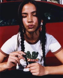 Close-up portrait of a young woman with braided dark hair wearing a graphic t-shirt in front of a red car, photographed by Ricky Alvarez.