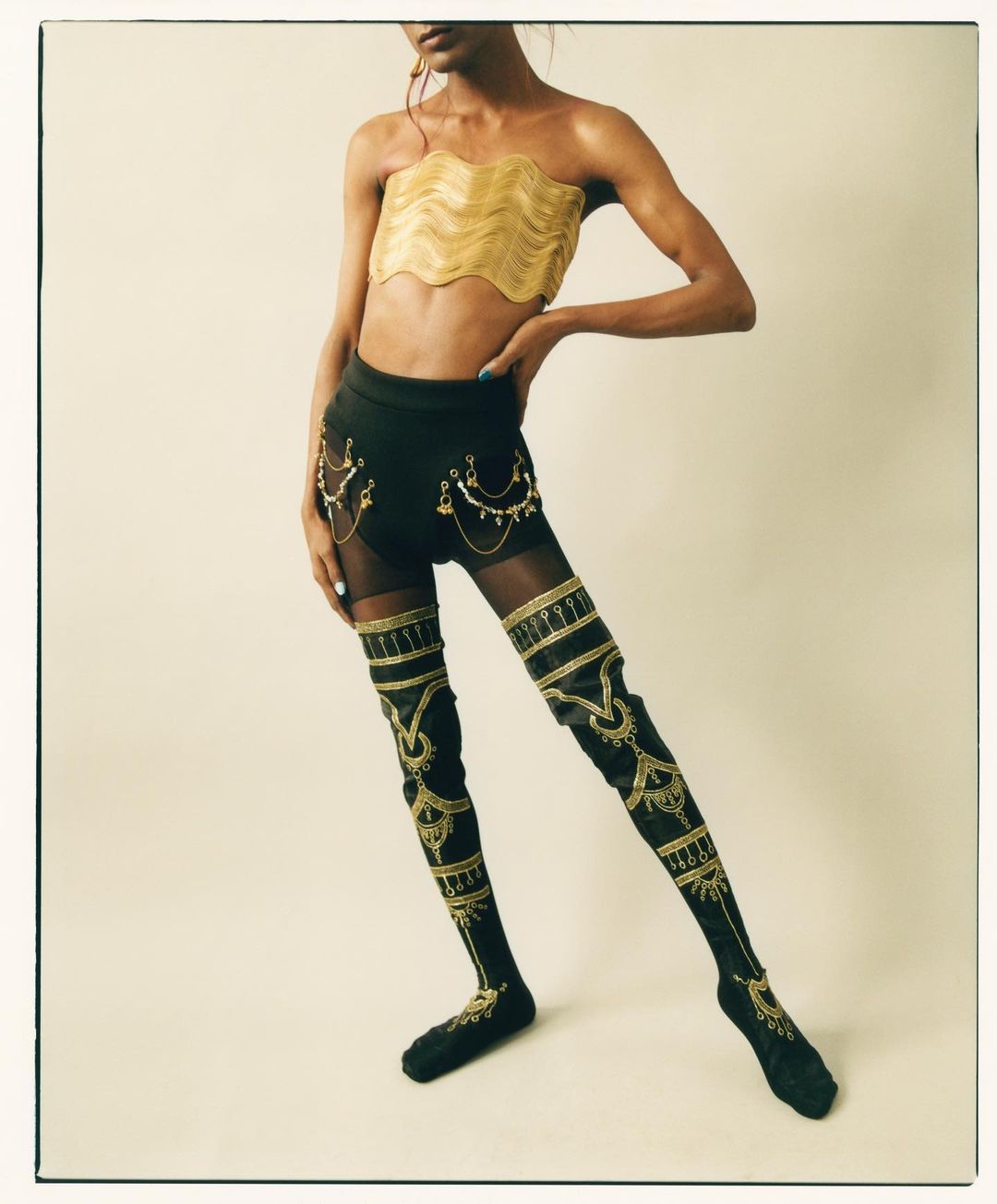 Person posing in gold tube top, shorts with chains, and high socks with gold accents in a studio with a cream background.
