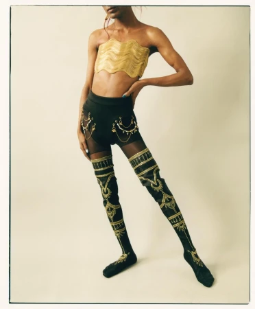 Person posing in gold tube top, shorts with chains, and high socks with gold accents in a studio with a cream background.