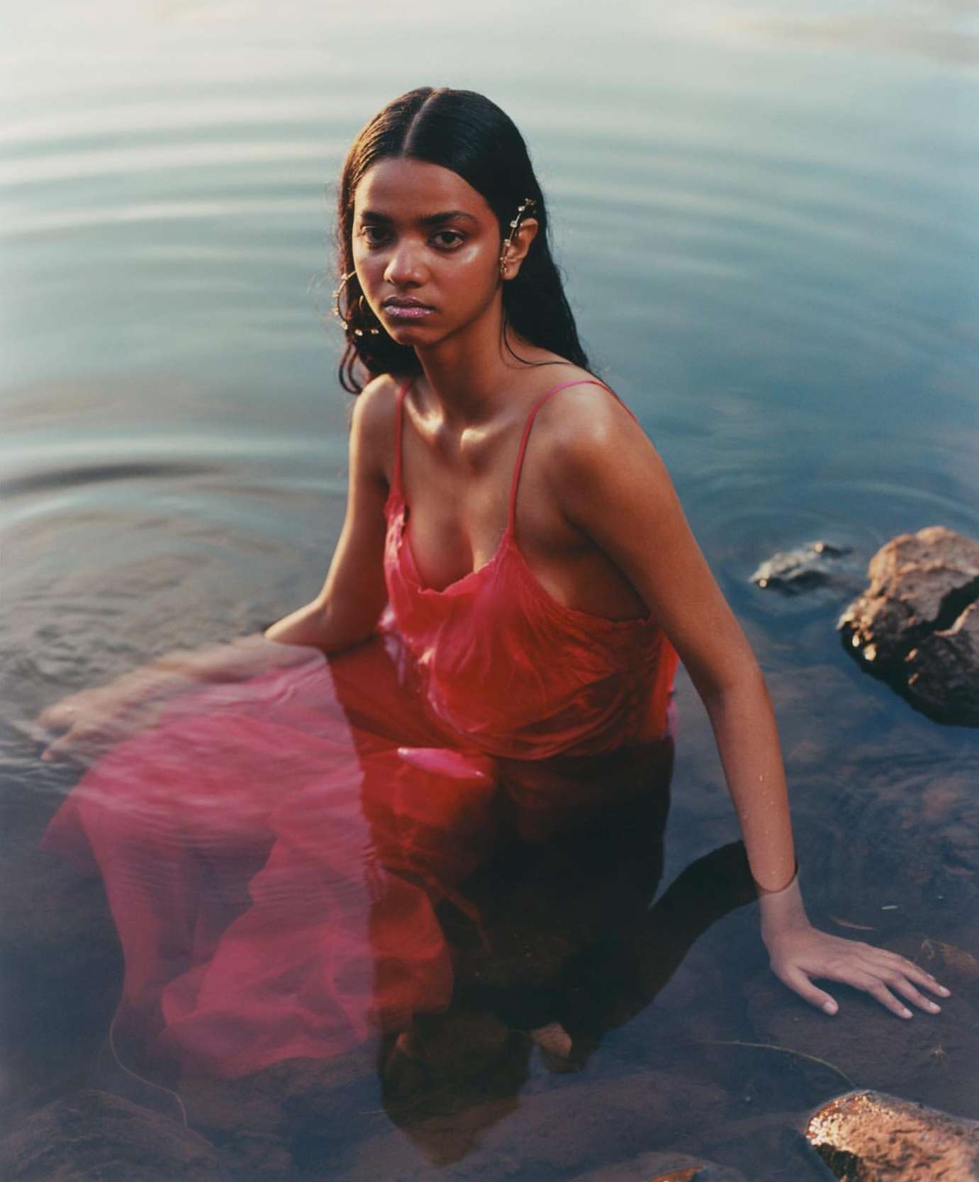 Woman in a red dress sits in water with rocks. Sunlight illuminates her skin and reflects off the water on a serene day.