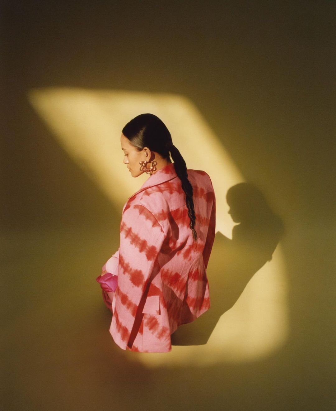 Woman in pink tie-dye blazer and earrings kneeling in a yellow-green studio with soft lighting and shadow, looking away from the camera.