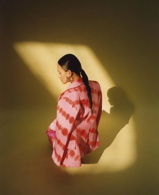 Woman in pink tie-dye blazer and earrings kneeling in a yellow-green studio with soft lighting and shadow, looking away from the camera.