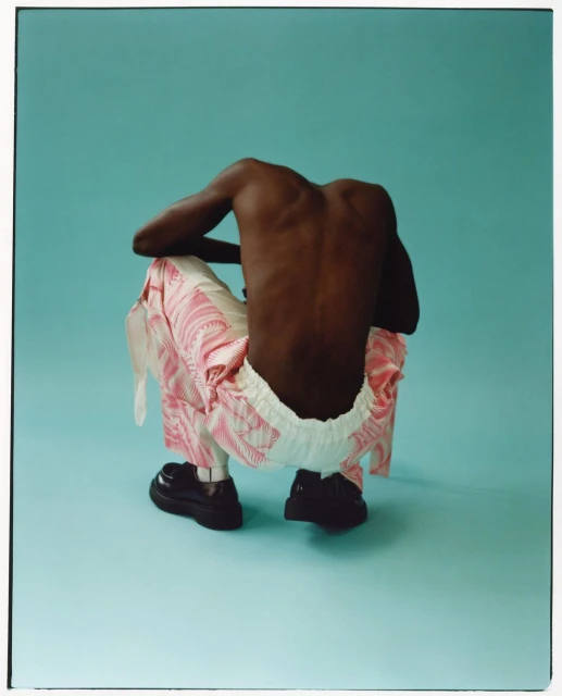 Rear view of a Black male crouching on a blue backdrop, wearing patterned pants and black shoes in a fashion editorial shot.