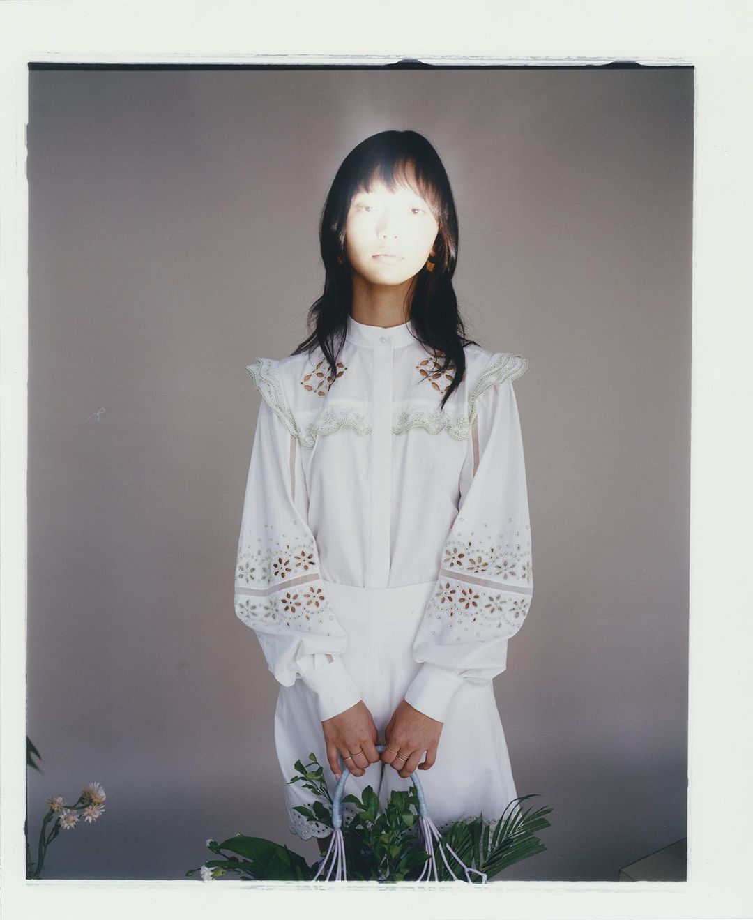 Asian woman in white vintage dress holding a floral arrangement against a grey backdrop with natural lighting and a photo studio environment.