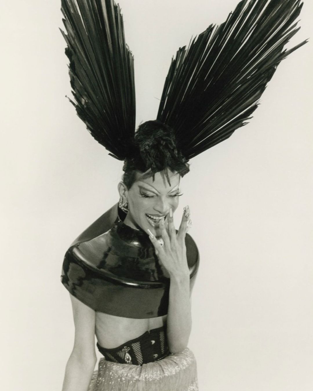 Stylish individual with large feather headpiece, metallic top, and jewelry against a white backdrop in a studio setting.
