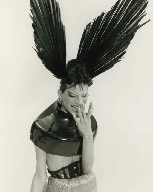 Stylish individual with large feather headpiece, metallic top, and jewelry against a white backdrop in a studio setting.