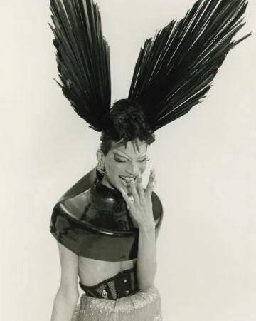 Stylish individual with large feather headpiece, metallic top, and jewelry against a white backdrop in a studio setting.