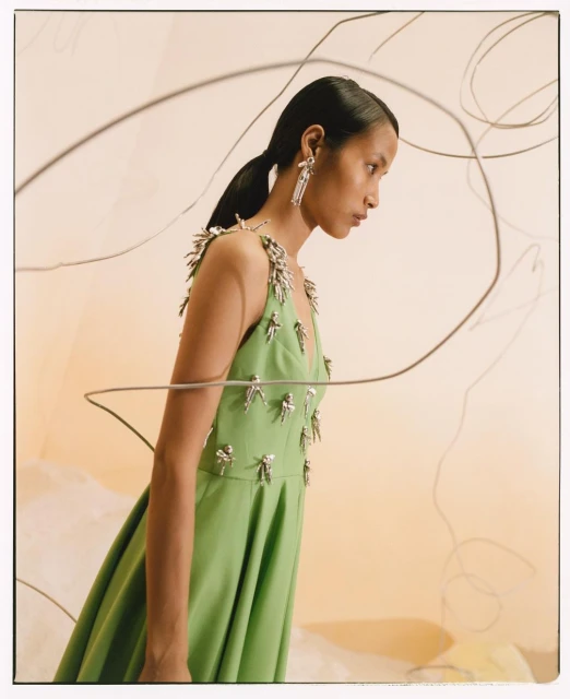 A woman poses in a green dress with silver accents against a light-colored background with wire art in a studio setting.