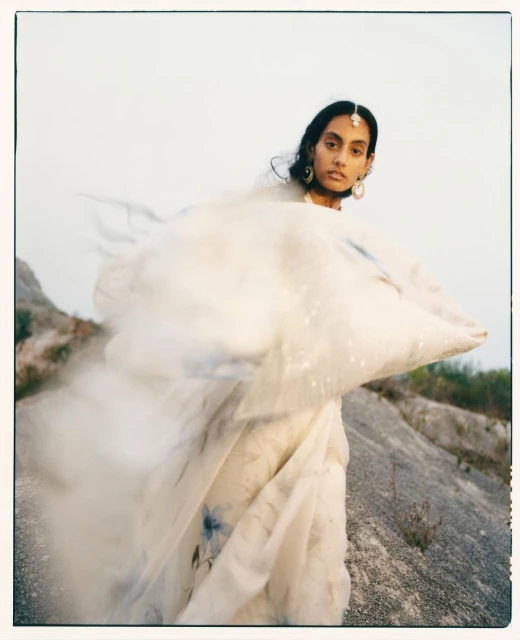 Woman in flowing gown poses outdoors on rocky terrain, wearing traditional jewelry and soft makeup in a fashion editorial style.