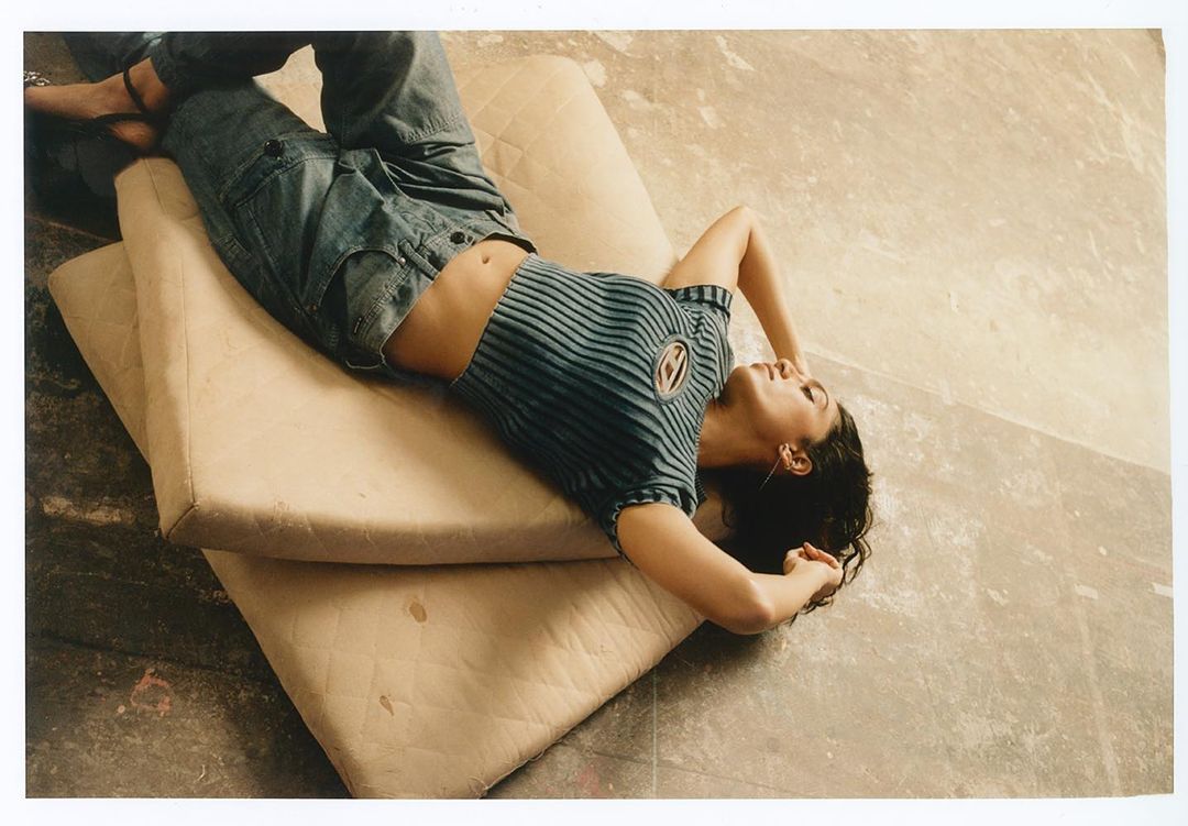 Woman in a blue striped top and jeans lounges on cushions indoors, eyes closed, capturing a relaxed and stylish moment.