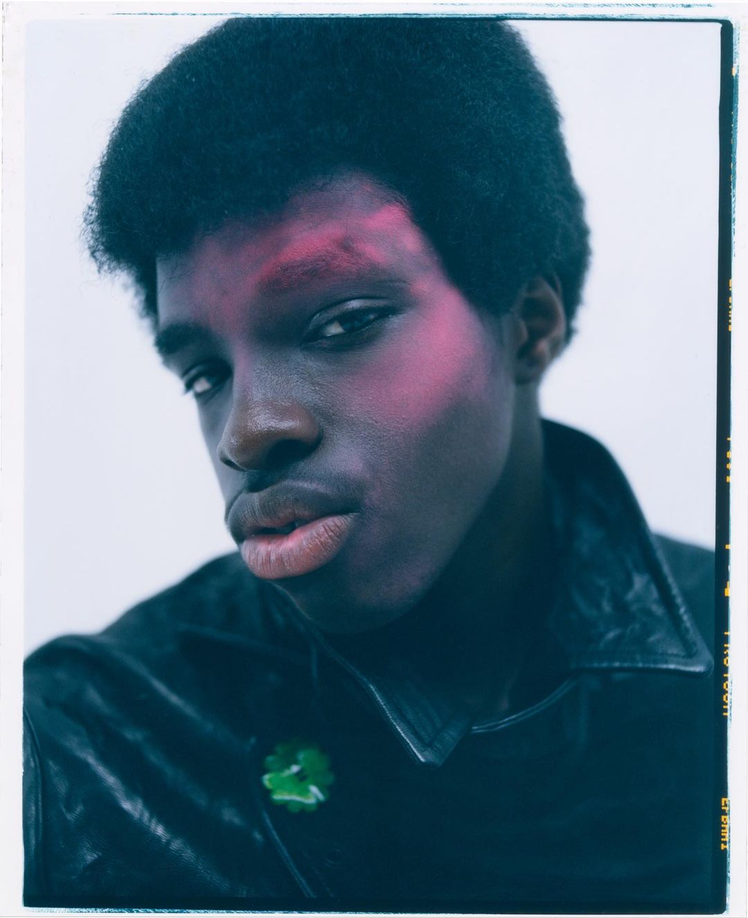 Close-up portrait of a Black adult male with red blush makeup on his face, wearing a black leather jacket against a light gray background.