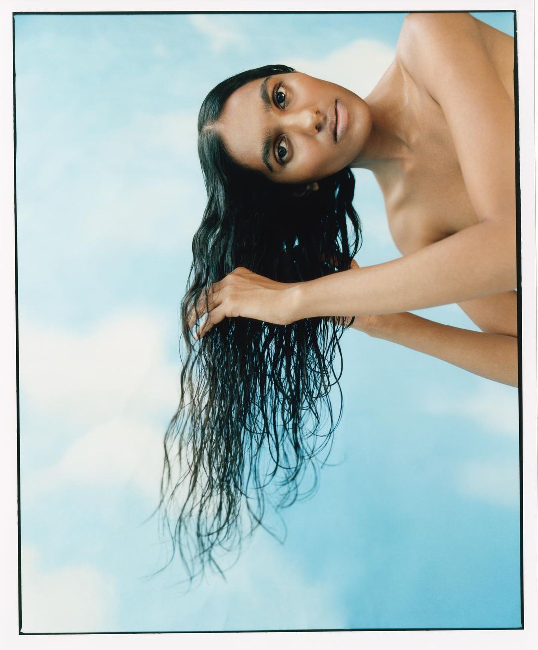 Adult woman with dark hair poses against a sky-blue cloud background, displaying beauty and style in a close-up studio shot.