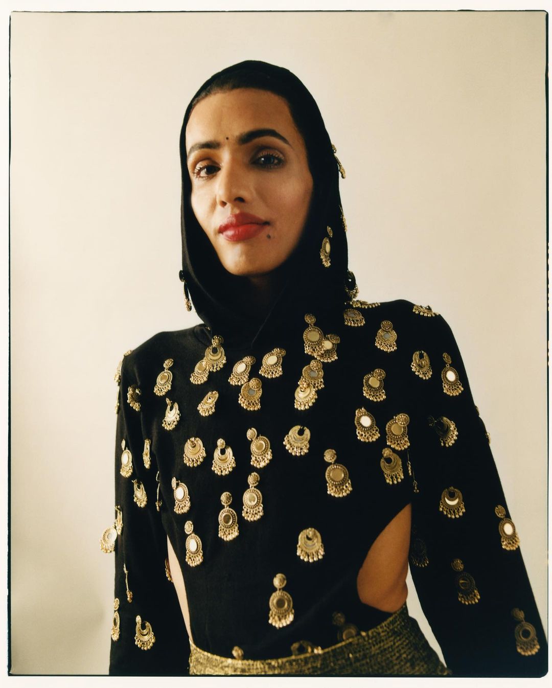 A person with dark hair and neutral makeup is wearing a black high-neck dress with gold embellishments against a light background.