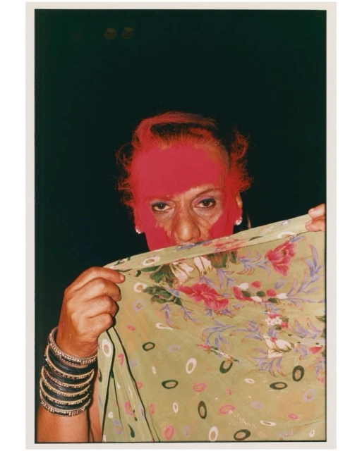 Portrait of an adult woman with pink powder on her face, partially veiled by a floral saree against a dark background, close-up shot.