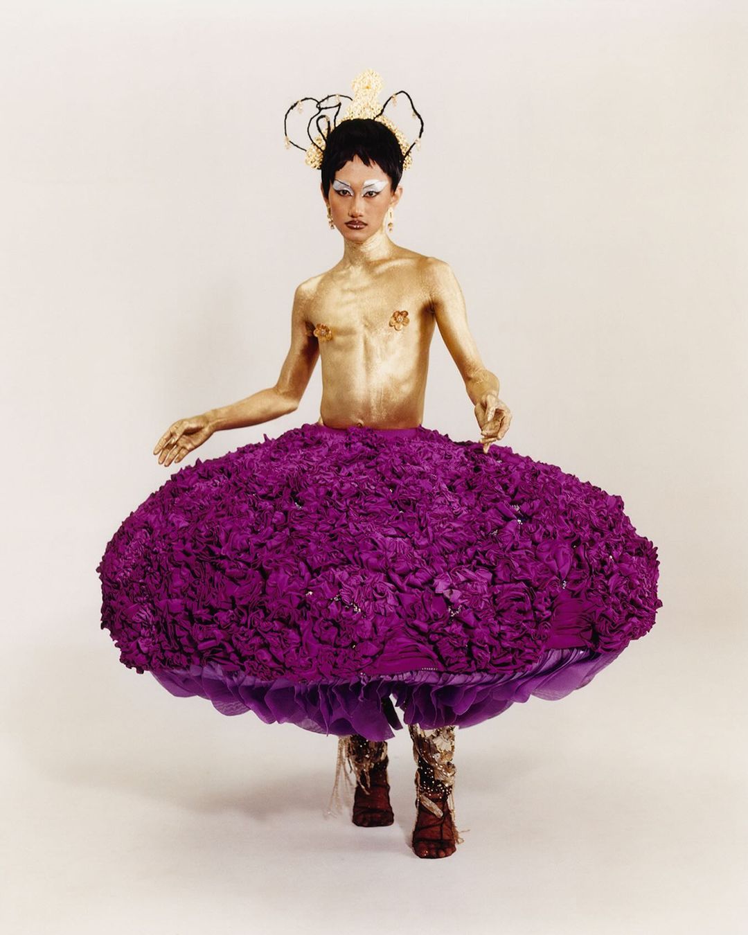 Glamorous individual with gold painted body, purple floral skirt, and ornate headpiece posing in front of a white backdrop in a studio shot.